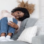 Loneliness. Depressed Black Lady Sitting On Couch With Smartphone, Loking At Screen